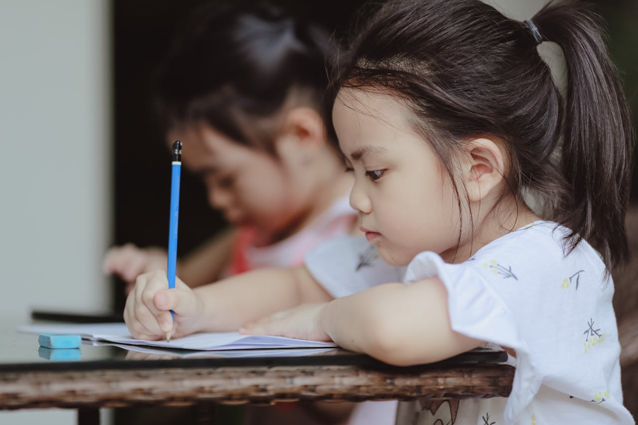 Adorable focused little Asian preschool girls sitting at table and doing tasks in copybooks in classroom